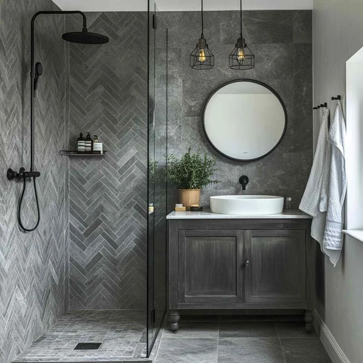 Dark gray herringbone shower tile with round mirror and vessel sink vanity